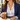 Woman in a white button-up shirt and navy blazer wearing the gold paper clip chain toggle necklace with a pearl drop, holding an iced coffee at a café table