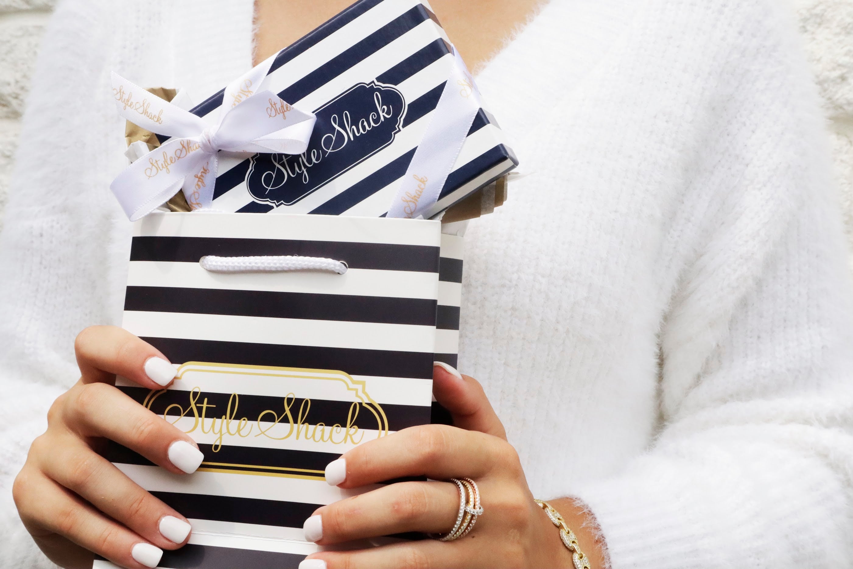 A girl in a soft white sweater holding a striped gift bag and striped box wrapped in a bow branded with Style Shack logos